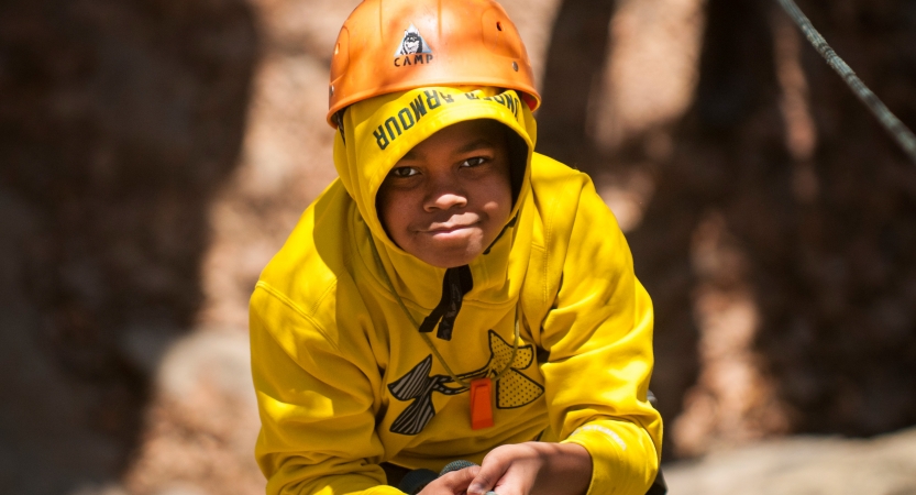 A person wearing a helmet is secured by safety gear as they look up at the camera while rock climbing